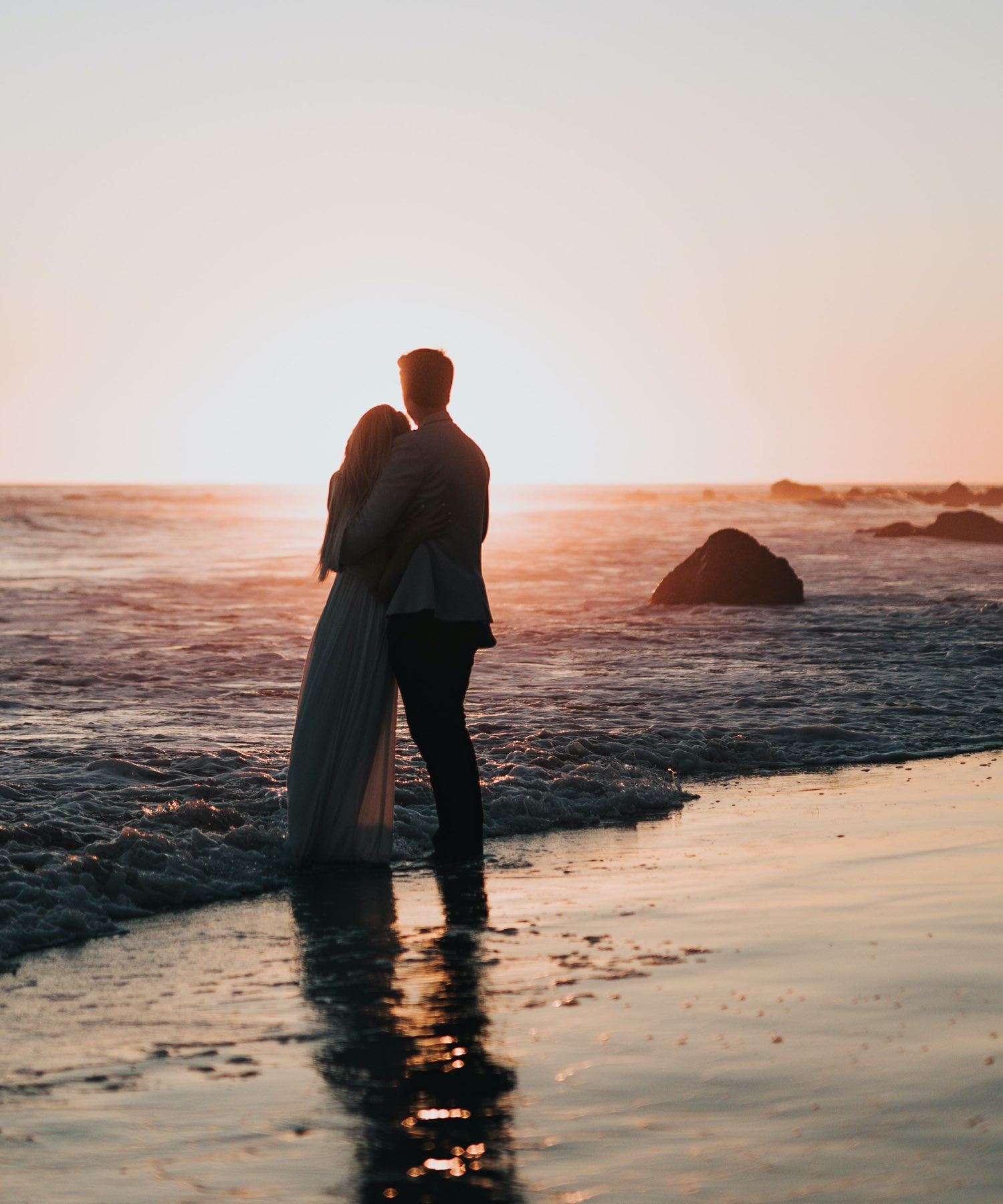 Сouple wearing a white gown and a black suit on the beach. Mobile image